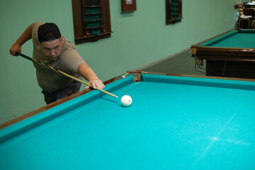Fototapeta premium A serious man holding a cue in his hands aims at a billiard ball. A man plays Russian billiards with his friends while having fun. A man is intently aiming at a ball on a billiard table.