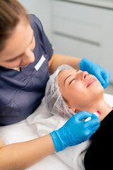 beautician doctor in gloves holding a brush and applying a health mask on the face and neck of a female client in a beauty salon