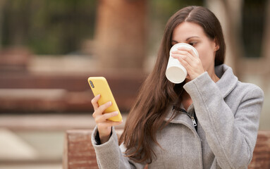 Long-haired brunette businesswoman checks her smartphone while drinking coffee sitting on park bench