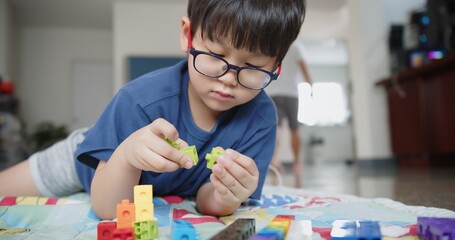 Portrait attractive smart cute Asian child boy wearing eyeglasses enjoy playing colorful tower blocks in living room