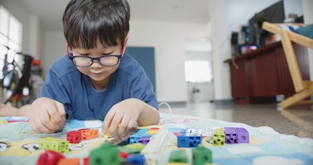 Portrait attractive smart cute Asian child boy wearing eyeglasses enjoy playing colorful tower blocks in living room