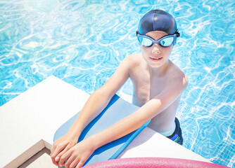 Active child (boy) in cap, sport goggles ready to learns professional swimming with pool board and swim noodles in swimming pool. Kid enjoying water. Healthy lifestyle.