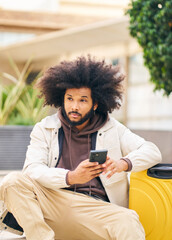 Young man with curly afro hair in the street sitting with the phone in his hand looking around him