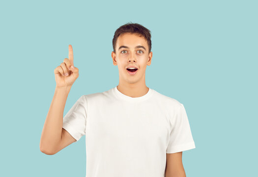 Clever young man or teen student boy in white T shirt isolated on blue background has genius idea, finds solution, solves mystery, points finger up, looks at camera with happy, amazed face expression