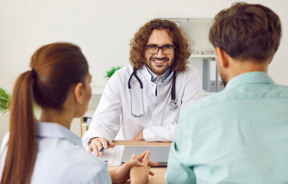 Smiling Happy Male Family Doctor Sitting At The Desk On Workplace And Consulting A Young Couple Husband And His Wife Sitting Back In Medical Clinic Planning Pregnancy. Health Care Concept.