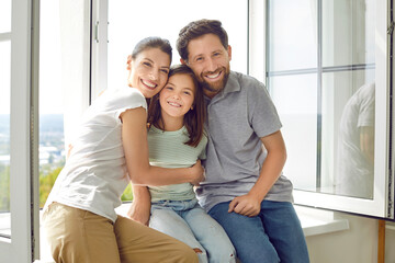 Portrait of smiling young happy family of three with daughter sitting on the windowsill at home and...