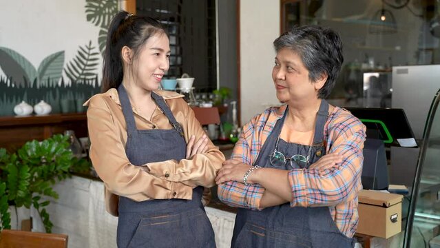 Senior mother and her Asian daughter Standing in front of the checkout counter taking orders in a coffee shop that is its own bakery. Standing with arms folded, looking at the camera, taking photos pr