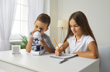 Two school children use microscope and look at microorganisms at magnification. Little boy studying something under microscope while little girl is taking notes on notepad for scientific hobby project
