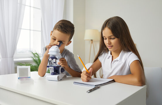 Two school children use microscope and look at microorganisms at magnification. Little boy studying something under microscope while little girl is taking notes on notepad for scientific hobby project