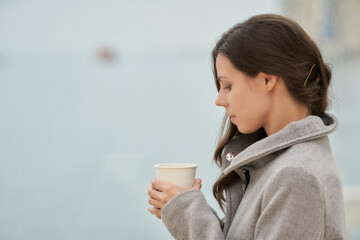 Young woman in a cozy coat holding a coffee cup, looking contemplative against a blurred background.