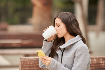 Young businesswoman drinking coffee while holding her smartphone sitting in the park.