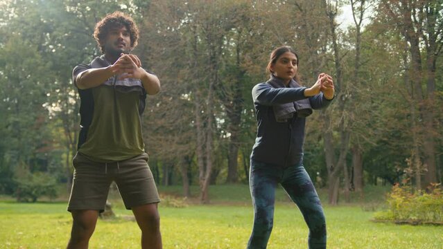 Active sporty two couple doing squats workout exercise in park nature outdoors sport together strong Arabian man guy and Indian woman girl athletes male female healthy lifestyle fitness squatting