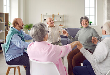 Group of happy senior people having fun in a retirement home, eldercare facility or community center. Cheerful retired old men and women sitting in a circle and holding hands - obrazy, fototapety, plakaty