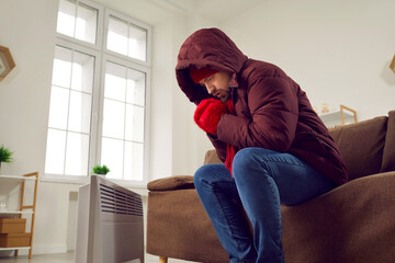 Unhappy young man sitting on couch feeling cold at home. Shivering man wearing winter jacket,...