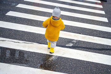 Stylish kid toddler girl in yellow rubber boots and coat on a striped pedestrian crossing on the...