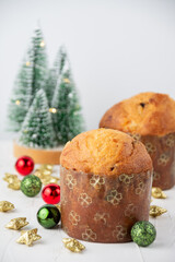View of two small Panettones on white board and table with illuminated trees, red and green Christmas decorations, white background, selective focus, vertical, with copy space