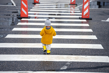 Stylish kid toddler girl in yellow rubber boots and coat on a striped pedestrian crossing on the road alone