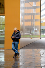 A young man stands with a smartphone near business buildings, cold weather