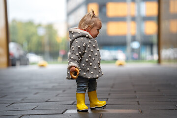 A cute stylish two-year-old toddler girl kid in a gray coat and yellow boots walks outdoors in the city in autumn season 