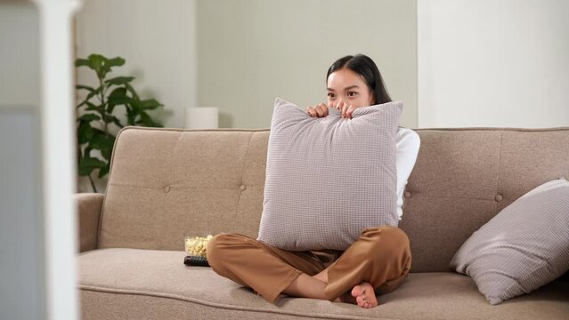 Young asian woman watching scary movie TV on sofa something excited in the film, looking and afraid emotion at living room, Mass media and emotion concept