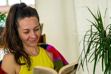 Naklejka premium Comfortable Literature Time: Woman Smiling and Reading a Book Wrapped in a Blanket at Home, Plenty of Copy Space