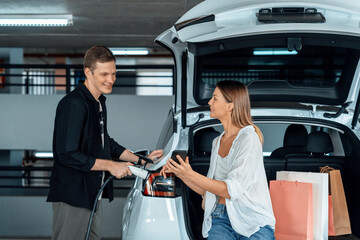 Young couple travel with EV electric car to shopping center parking lot charging in downtown city...