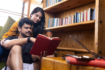 Young couple seating on couch at home or cafe or in library near window reading a book enjoying...