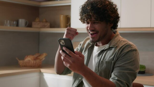 Satisfied ecstatic man receive job offer opportunity winning Indian happy Arabian male good news smartphone win. Excited guy winner celebrating success achievement mobile phone victory at home kitchen