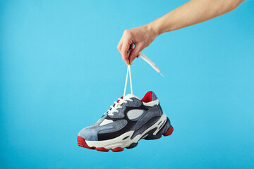 Hand holds hanging colored running sneakers by the laces on blue pastel background. Hand with a new...