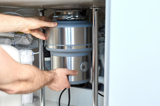 A plumber with gloved hands installs a household waste shredder for the kitchen sink. A technician installs or repairs a household waste shredder in the kitchen.