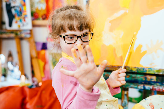 Cute little child girl with down syndrome giving high five to camera during painting at easel at art class. Kids mental development concept.