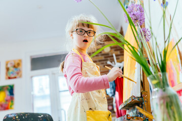 A talented little girl with Down syndrome is studying at an art school, dressed in an apron stained...