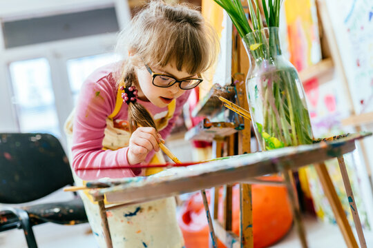 A talented little girl with Down syndrome is studying at an art school, dressed in an apron stained with paint and holding a brush in her hands. Development of children with special needs.