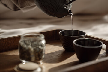 Black cast iron tea pot with herbal tea set up on wood tray on the bed. Traditional Asian Tea Set -...