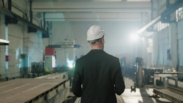 In front of the camera taking video from the back of main manager with safety helmet he holding the plan of work and standing in the middle of manufacturing production zone