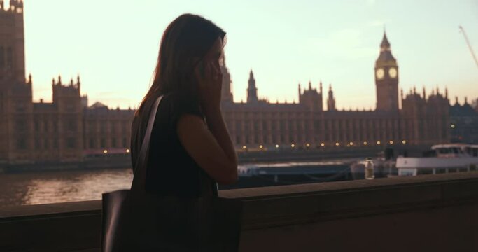 Happy local attractive brunette female talks via smartphone and smiles, while walking along the Thames river.