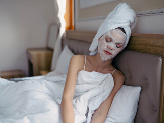 Young woman lying on bed with moisturizing beauty face mask and white towel on her head after shower, lifestyle home skincare, smile.