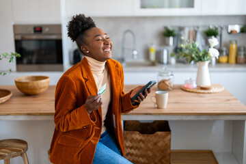 Enthusiastic charming black woman feeling excited holding mobile phone and credit card satisfied with online payment possibilities, rejoicing approved loan.