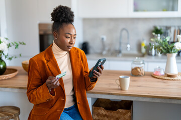 Serious businesswoman sitting at kitchen counter at home, holding credit card while looking at phone screen, paying bills online, using smart phone for electronic banking. - obrazy, fototapety, plakaty