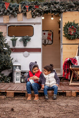 African American boy and girl are sitting at the camper on wooden pallets, playing with a Christmas snow globe. Brother and sister shake the ball, snow flies in it. Christmas holidays outside the city