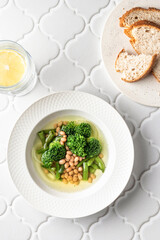 Vegetarian minestrone soup with green vegetables and beans served in white plate on white tile background, top view