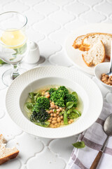 Vegetarian minestrone soup with green vegetables and beans served in white plate and bread on white tile background