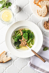 Vegetarian minestrone soup with green vegetables and beans served in white plate on white tile background, top view