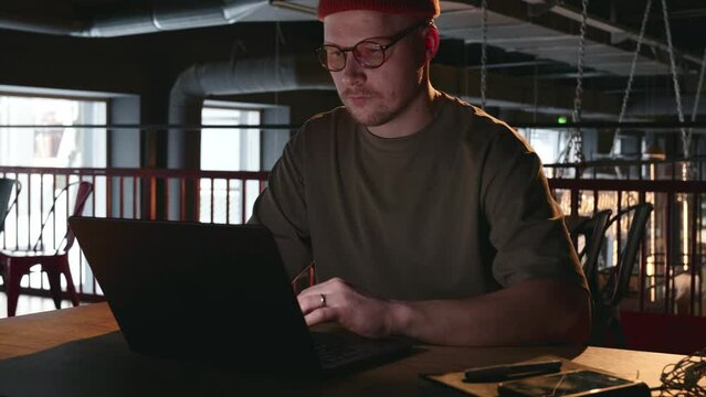A young millennial man in glasses and a fashionable hat is working on a laptop in a dark room of an empty loft. He moves closer to the table where the laptop is and starts working. High quality 4k