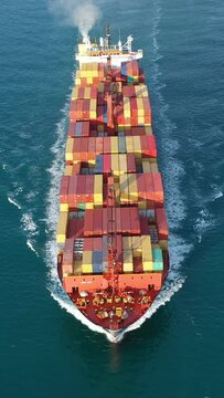 Container Ship underway calm waters with a dolphin surfing the bow wave. Vessel with white trails all around on nice deep green shade of ocean. Complete deck extends from prow to aft. Aerial vertical