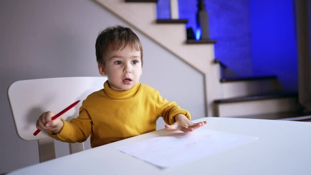 Small toddler in yellow sweater sitting at desk. Kid holds a red pen and waving it. Child drawing at home.