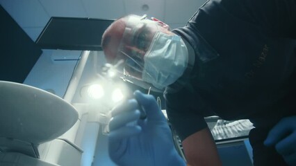 Male dentist in shield mask and latex gloves turning the light of dentist lamp on camera, spraying a stream of water from turbine, patient point of view. High quality 4k footage