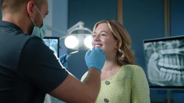 Young dental expert in medical gloves and mask examinating teeth of smiling blonde woman using tooth scraper, concave mirror standing in illuminated office. High quality 4k footage