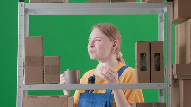 Frame on green background from chromakey. Young woman in working uniform. Depicts a storekeeper in a warehouse. She is holding a cup with a sandwich, eating and smiling, she is happy, she has lunch