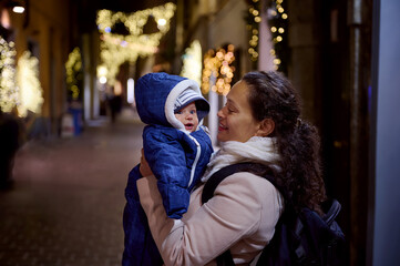 Cheerful happy young woman mother smiles holding her baby boy, strolling the street of elegant Como city at night
